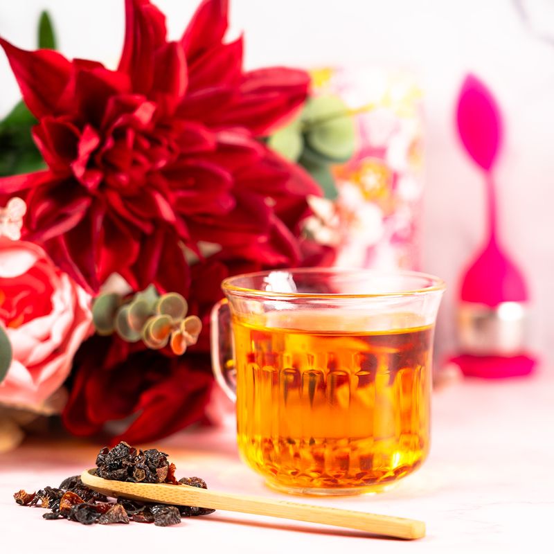 Cup of Organic Rosehip tea with dried rosehips on a wooden spoon and bright red flowers in the background.