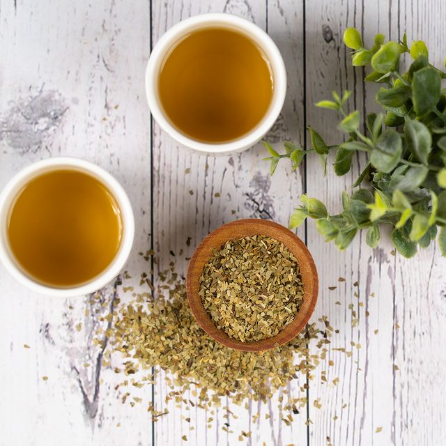 Two cups of Organic Yerba Mate tea beside a wooden bowl filled with loose yerba leaves, some scattered on a white rustic wooden surface with green foliage for decoration.