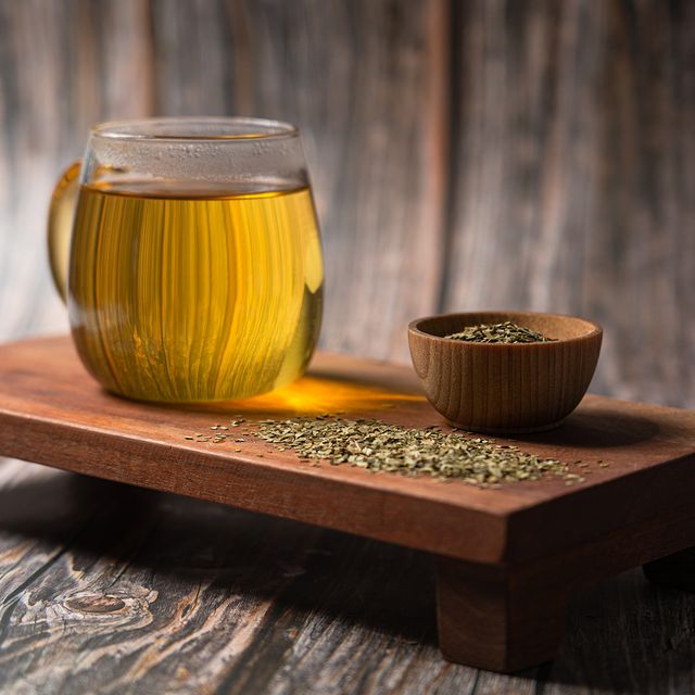 A glass mug of Yerba Mate tea with a warm golden hue placed on a wooden tray beside a small wooden bowl of loose yerba leaves, on a rustic wood surface.
